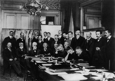 Marie Curie at the first Solvay Conference (1911), seated second from the right, conferring with Henri Poincaré, with notable scientists like Rutherford, Einstein, and Langevin standing nearby.