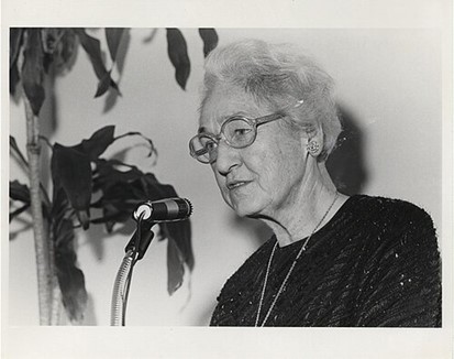 Dr. Virginia Apgar speaking at a podium during an event, wearing glasses and a dark outfit.