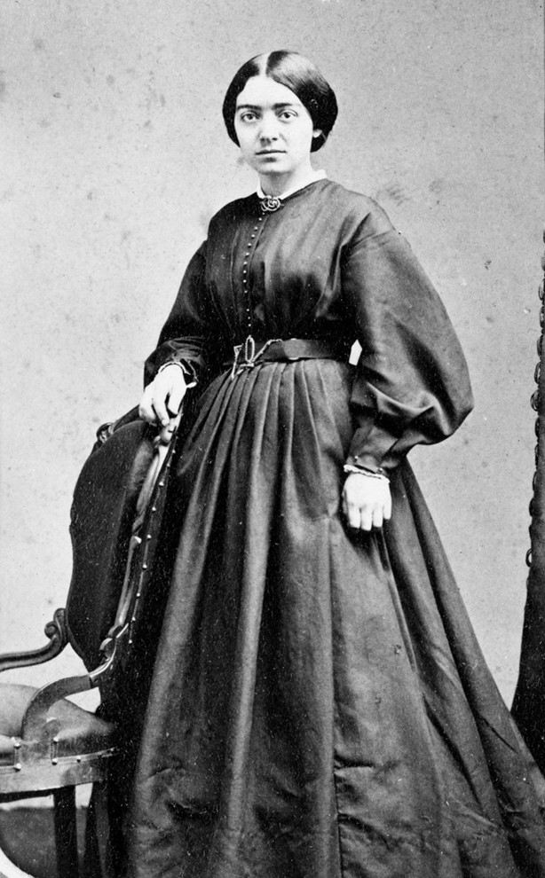 Mary Putnam Jacobi in a full-length portrait wearing 19th-century attire, standing beside a chair, showcasing her determined and intellectual demeanor.