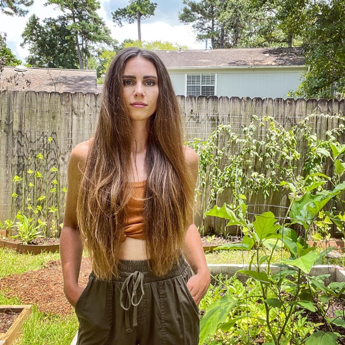 Jaelin’s daughter Bridget with long, flowing brown hair standing confidently in a lush garden, surrounded by greenery and sunlight.