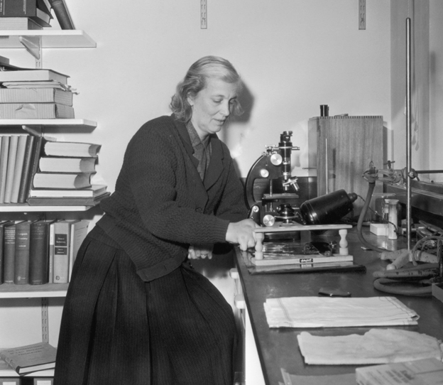 Black and white photo of Dorothy Crowfoot Hodgkin working with a microscope in her laboratory, surrounded by scientific equipment and books.