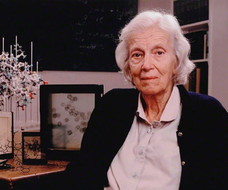 Elderly Dorothy Crowfoot Hodgkin in her later years, seated with molecular models and scientific diagrams in the background.